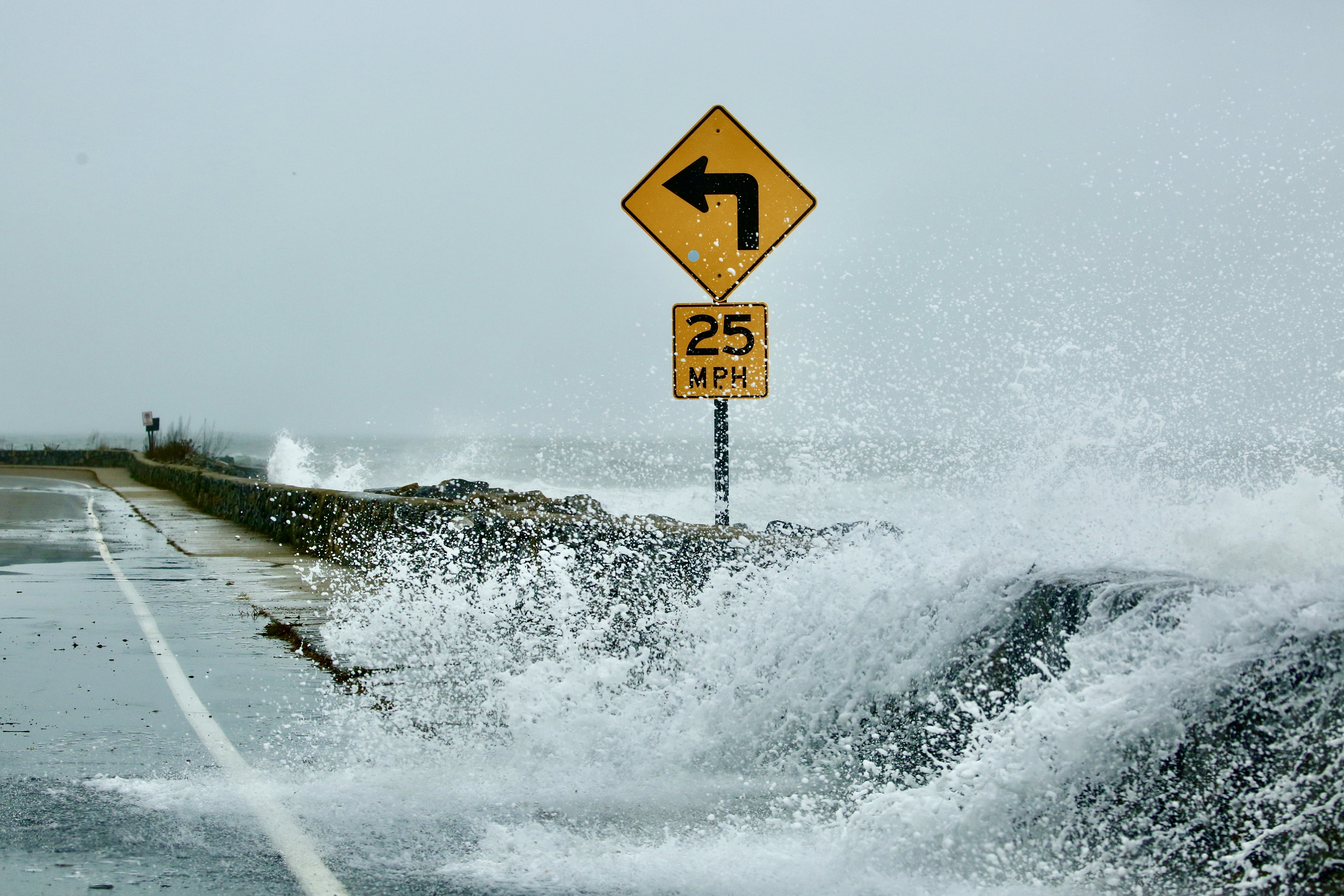 a wave breaks over a seawall and into a roadway.