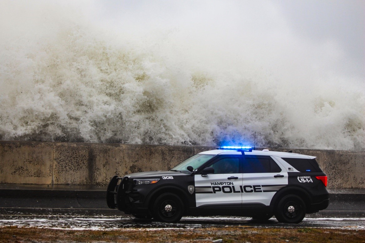 A Hampton police cruiser with its lights on sitting beside a sea wall with a giant wave crashing above it.