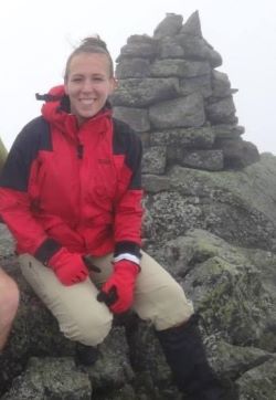 Smiling woman wearing a red and black coat sitting near cairn of rocks.