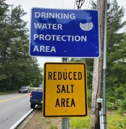 Blue street sign reading "Drinking Water Protection Area" above yellow sign "Reduced Salt Area."